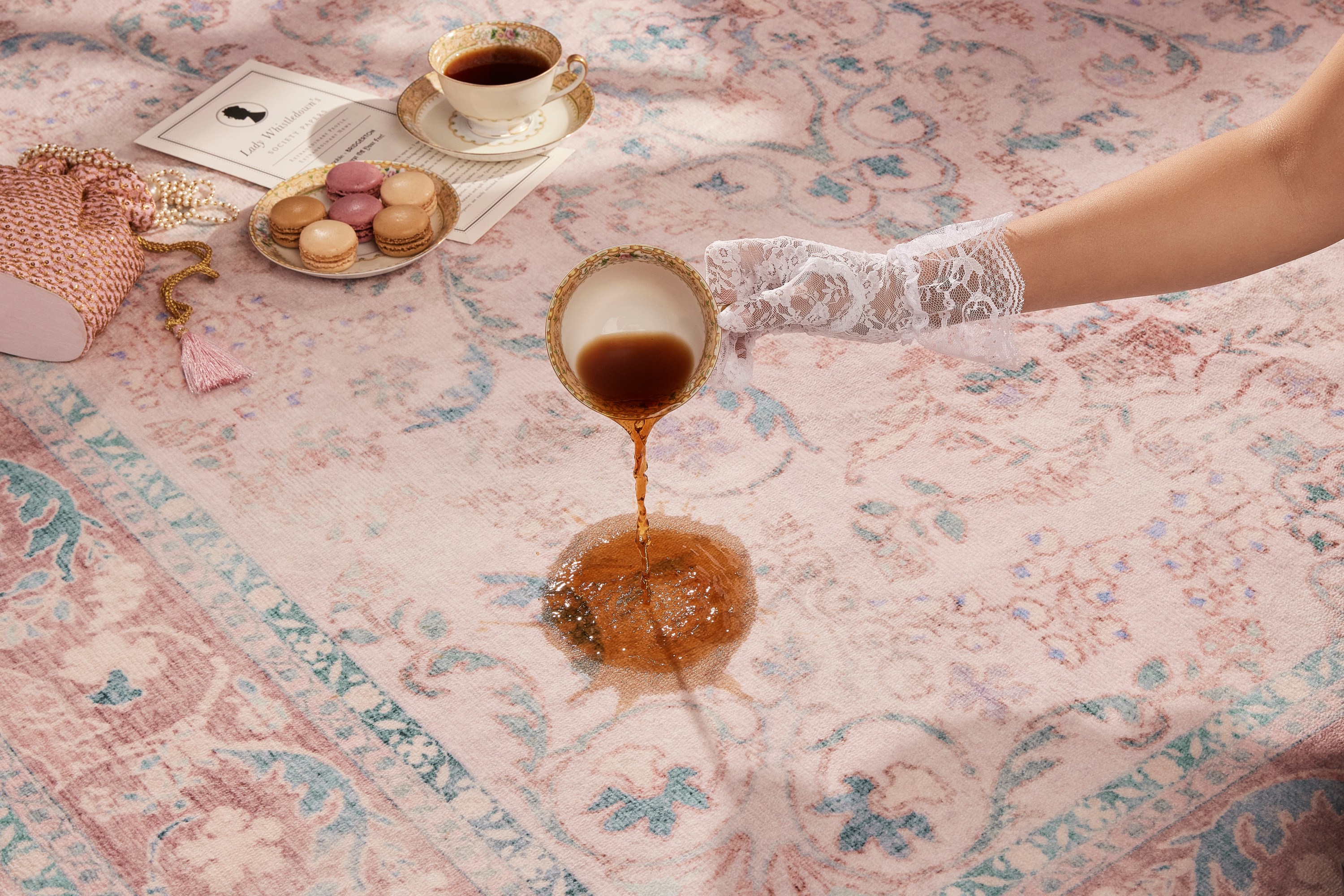 A laced glove hand spills tea on a blush pink rug.